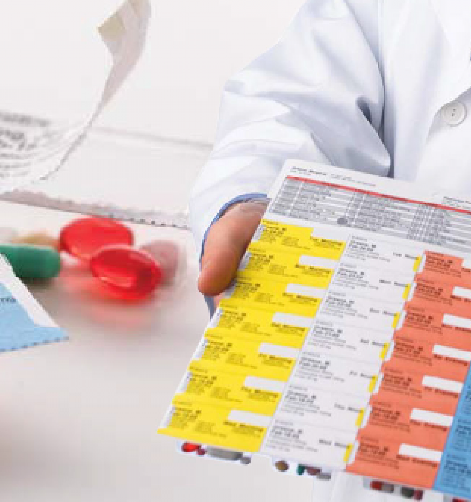 A person in a white coat is holding a color-coded medication schedule. There's a pill pack labeled 