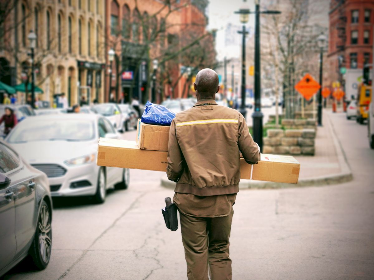 A delivery person is carrying packages while walking down a city street. Vehicles and buildings line the background.