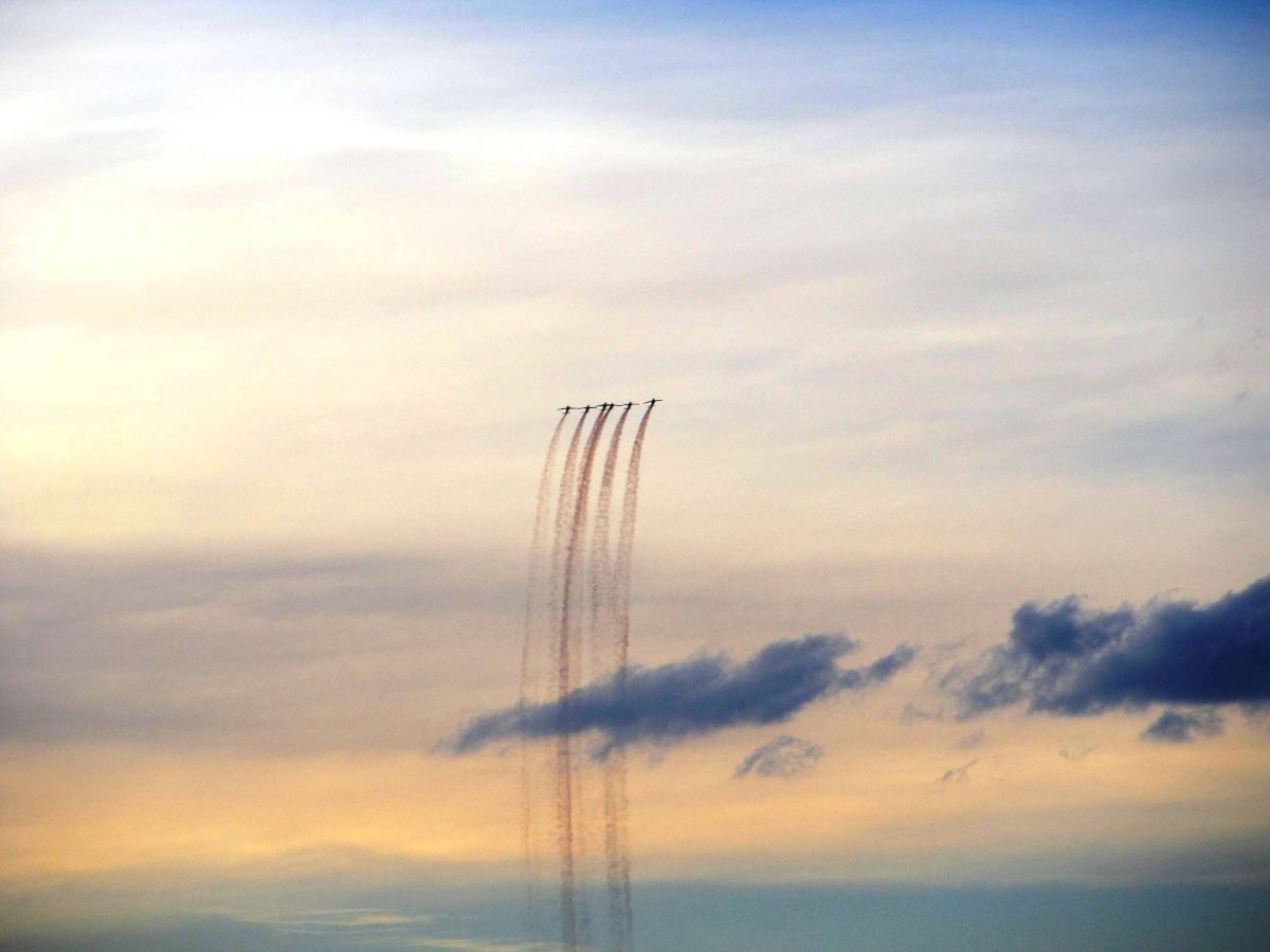 The image shows six airplanes flying in close formation with trails of smoke behind them against a backdrop of a cloudy, sunset sky.