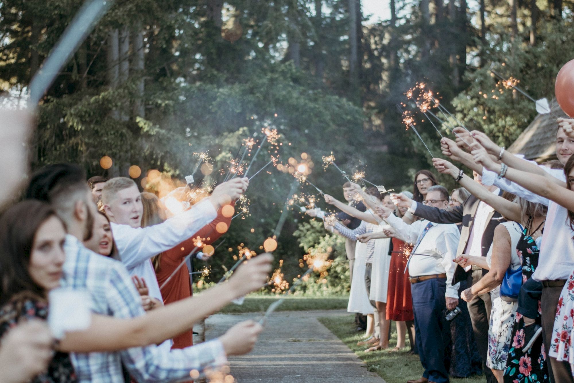 A group of people, likely at a celebration or wedding, are standing in two lines holding sparklers, creating a festive atmosphere.