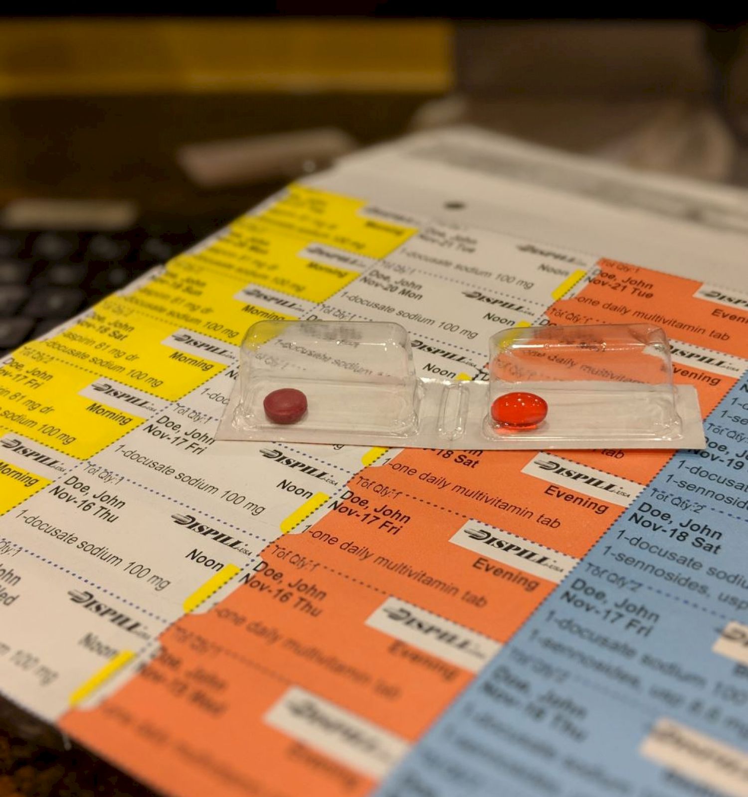 The image shows a color-coded medication schedule with two red pills in small plastic bags placed on top of it.