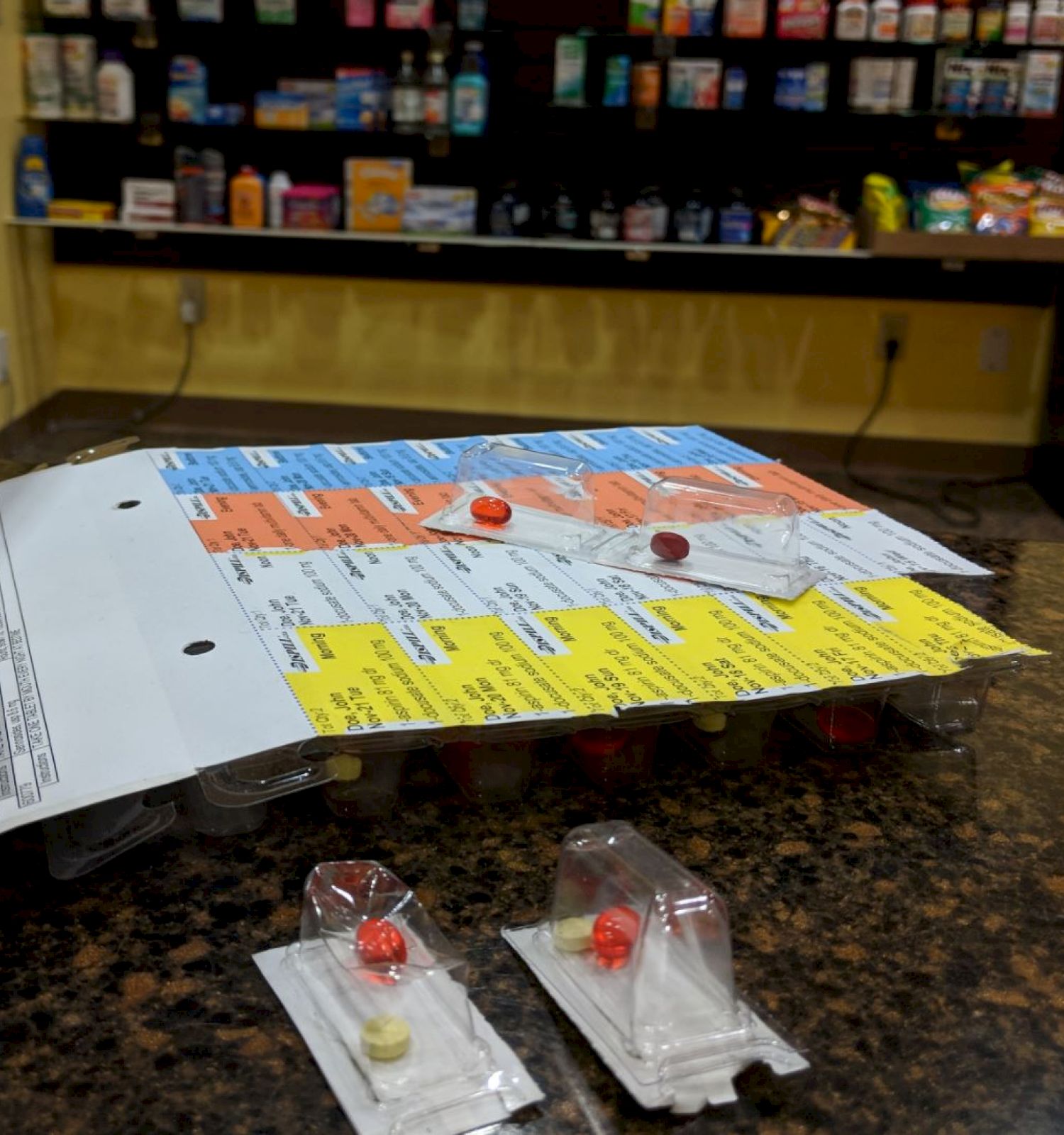 A close-up of medication blister packs on a countertop; a well-stocked pharmacy shelf is visible in the background.