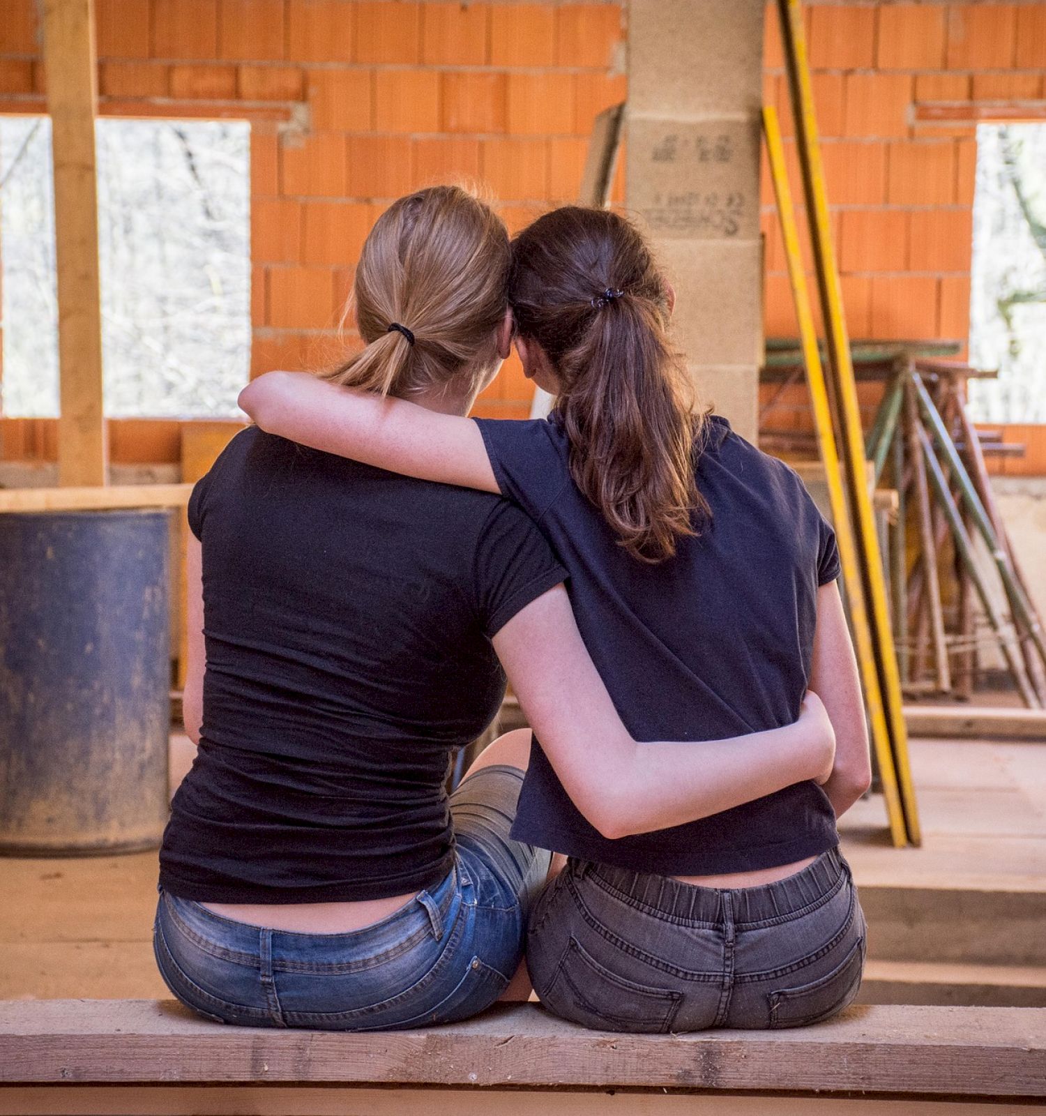 Two people with their arms around each other, sitting on a wooden plank in a construction site with unfinished walls and building materials.