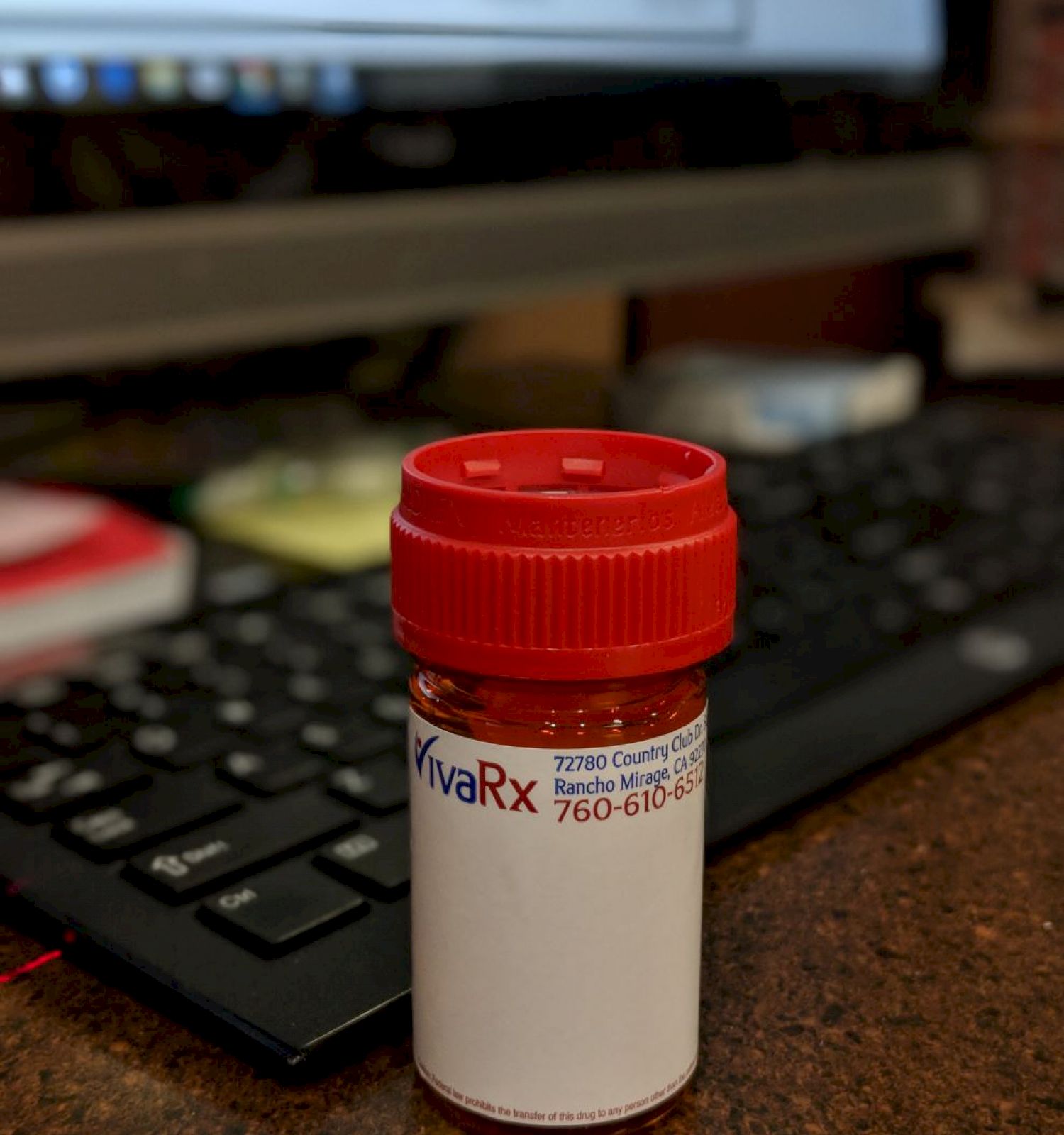 A small prescription bottle with a red cap is placed in front of a keyboard and computer screen on a desk.