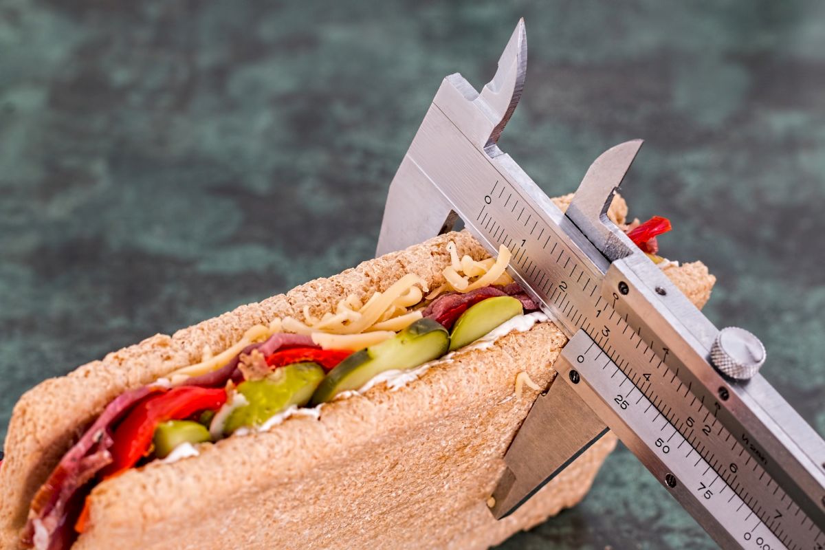 A sandwich with various fillings is being measured with a caliper against a green background.