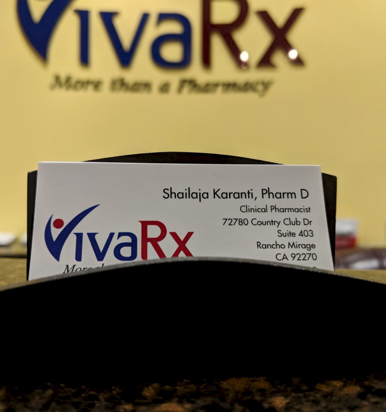 The image shows a business card holder with a card for VivaRx, displaying contact information for a clinical pharmacist. The background shows the VivaRx sign.