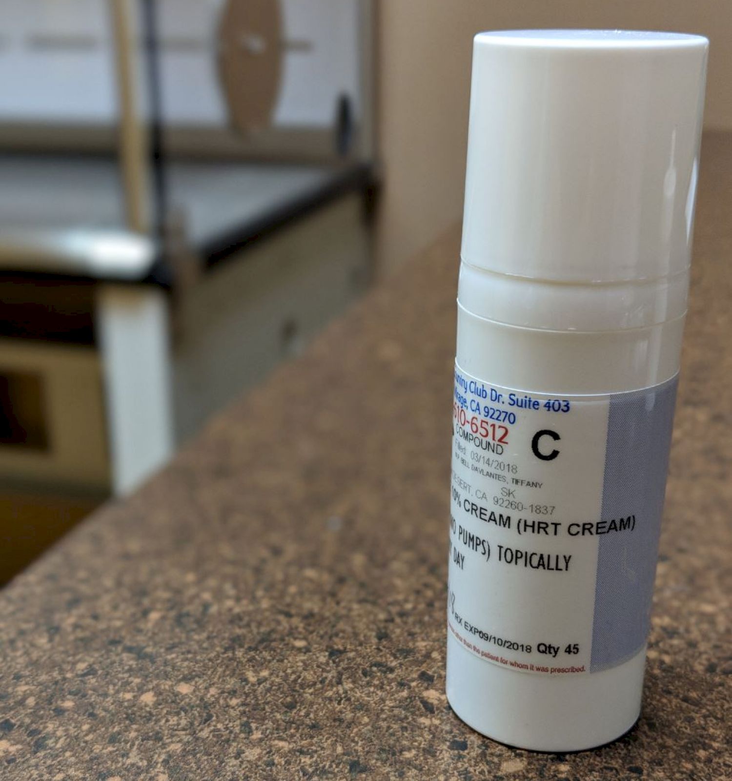 The image shows a white bottle with a medical label containing a cream, placed on a speckled countertop in what appears to be a clinical setting.