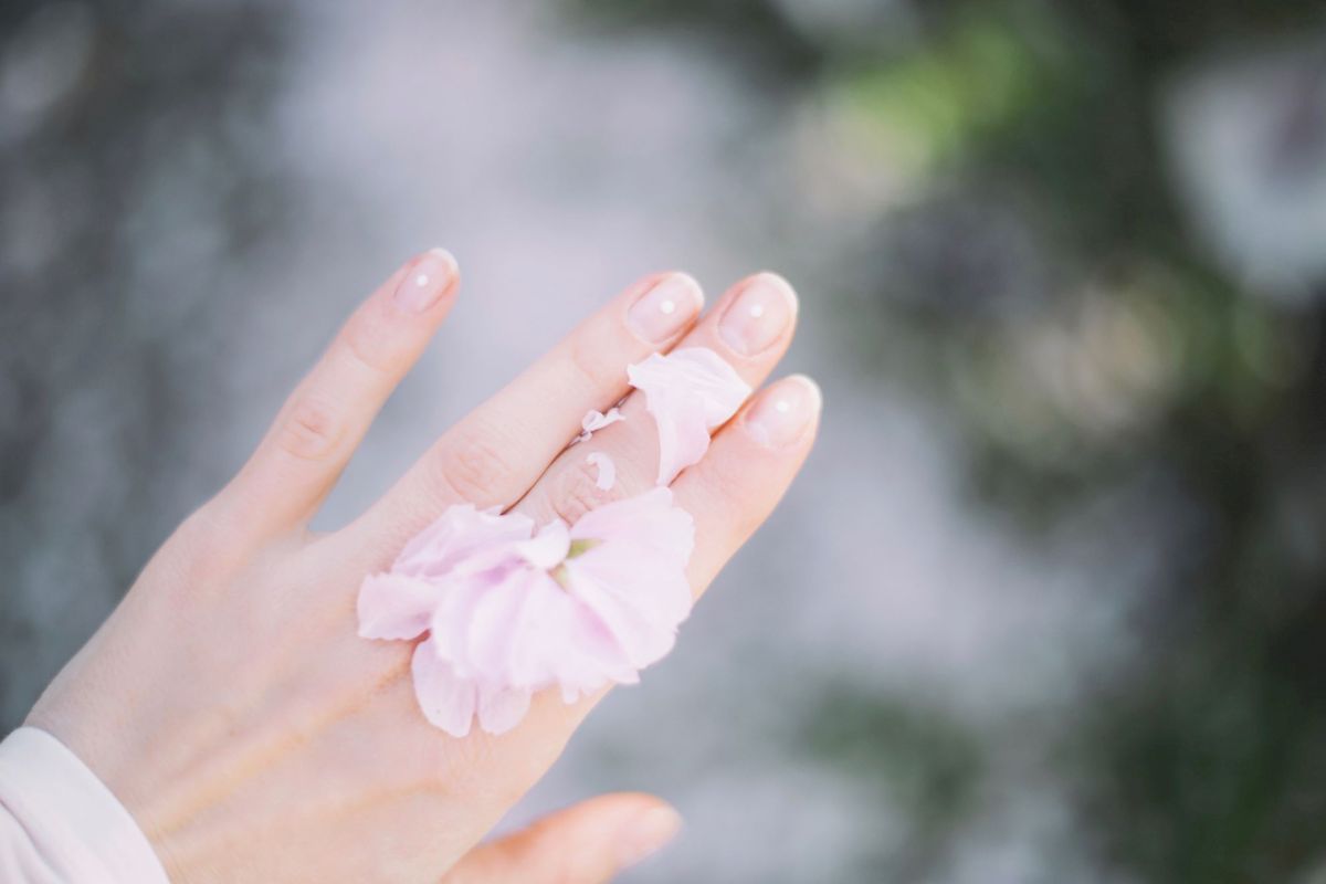 A delicate hand adorned with a soft pink flower rests against a blurred natural background, creating an ethereal and serene atmosphere.
