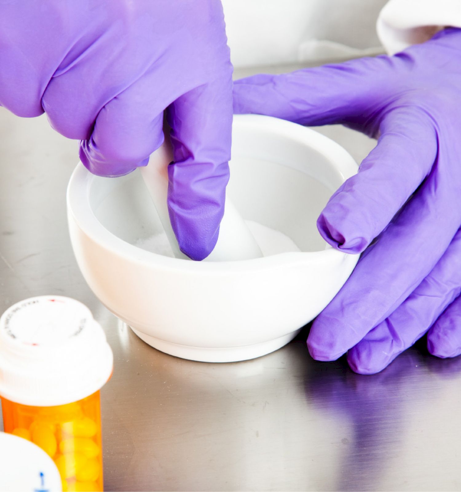 A person wearing purple gloves is using a mortar and pestle, with prescription pill bottles placed nearby on a stainless steel surface.