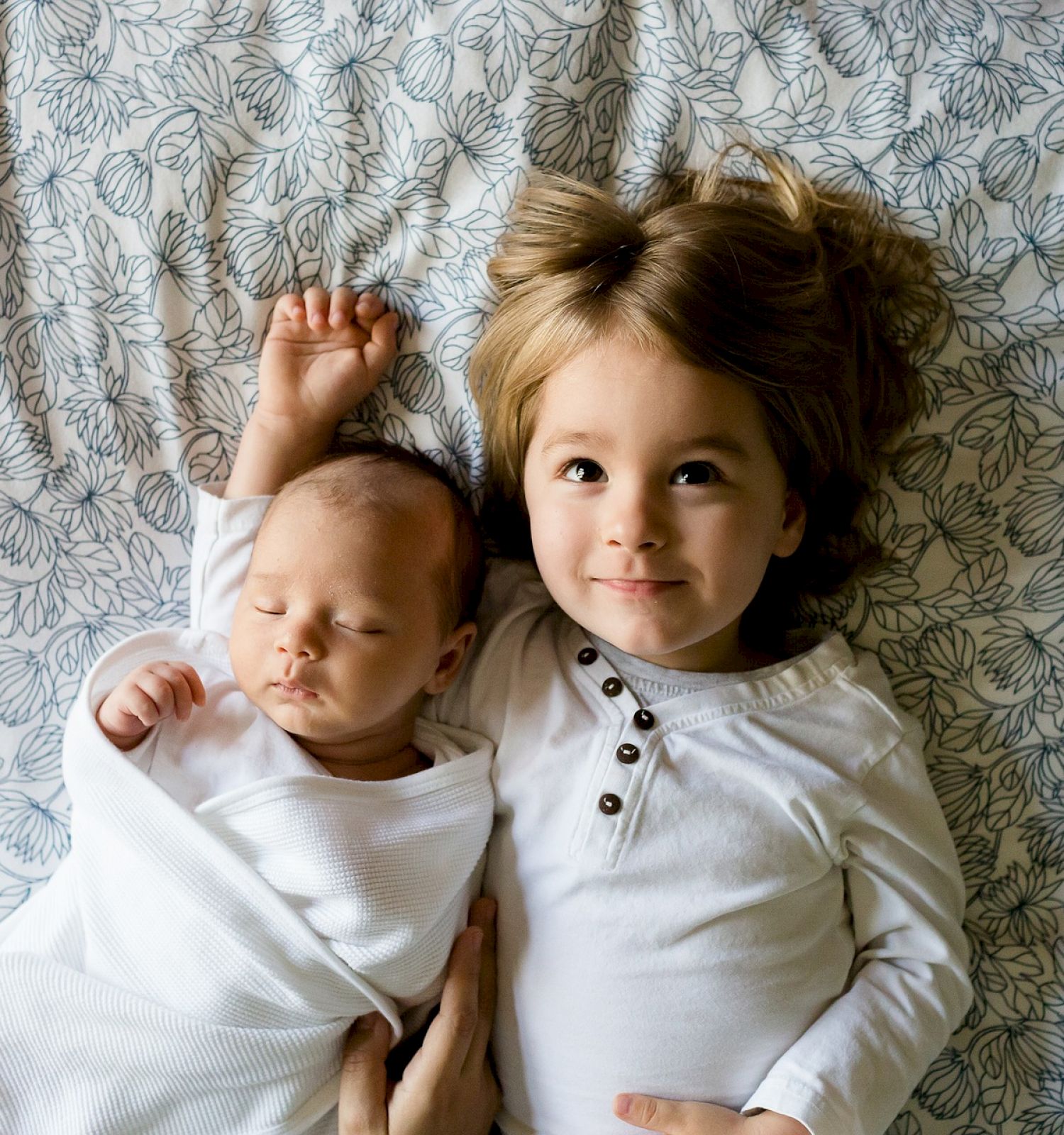 A baby wrapped in a white blanket lies next to an older child wearing a white shirt on a bed with a floral-patterned sheet.