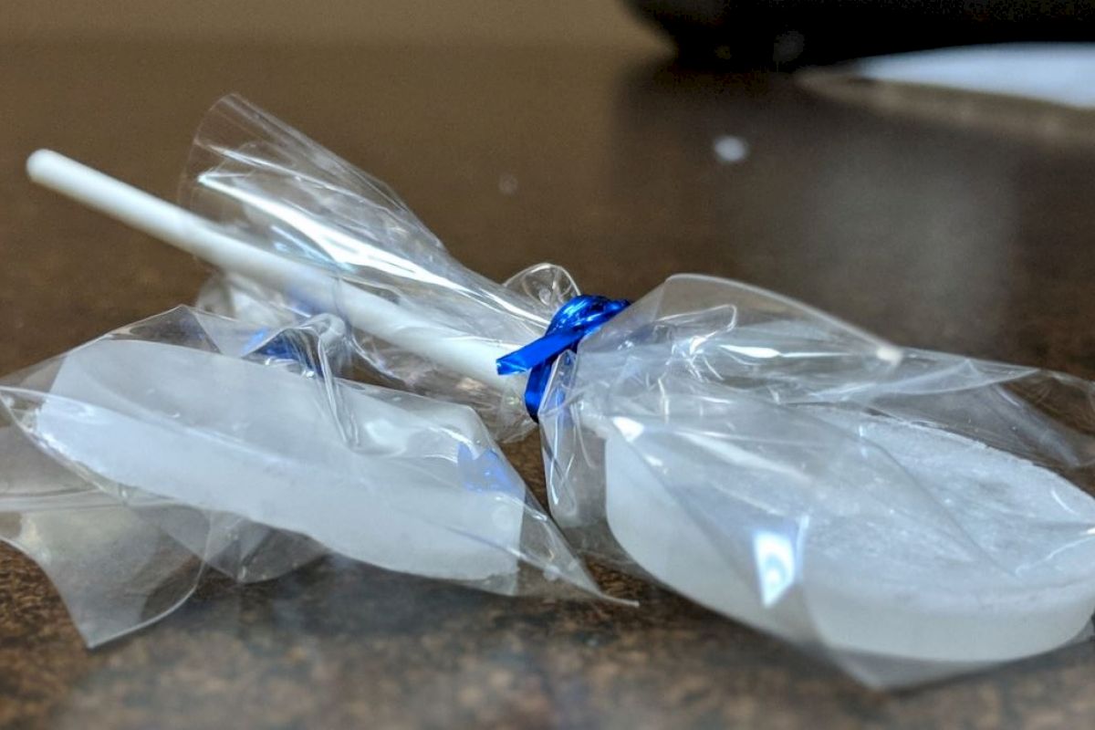 The image shows a white lollipop wrapped in clear plastic with a blue twist tie, placed on a dark surface.