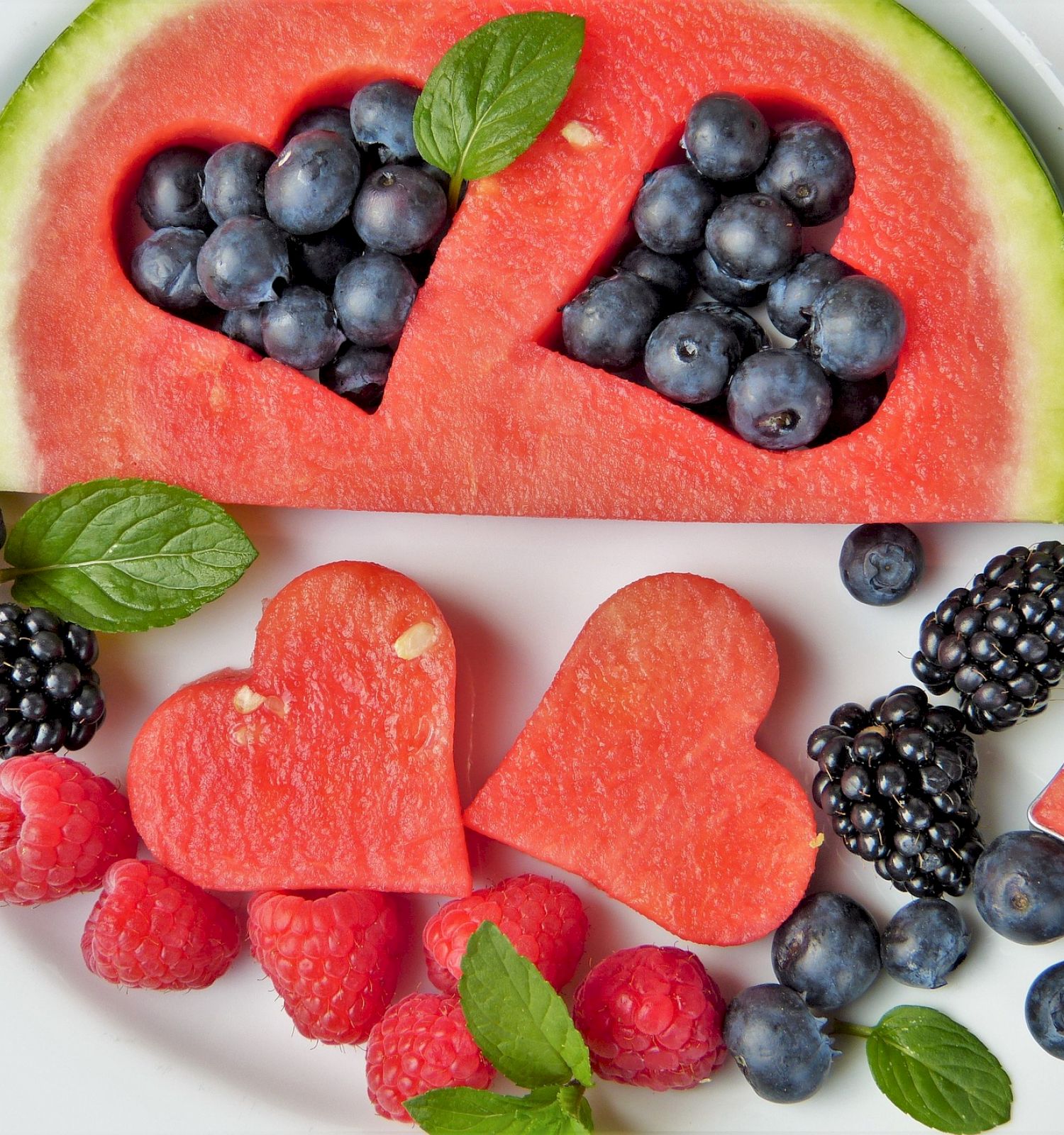 A plate with watermelon hearts, blueberries, raspberries, blackberries, and mint leaves arranged decoratively.