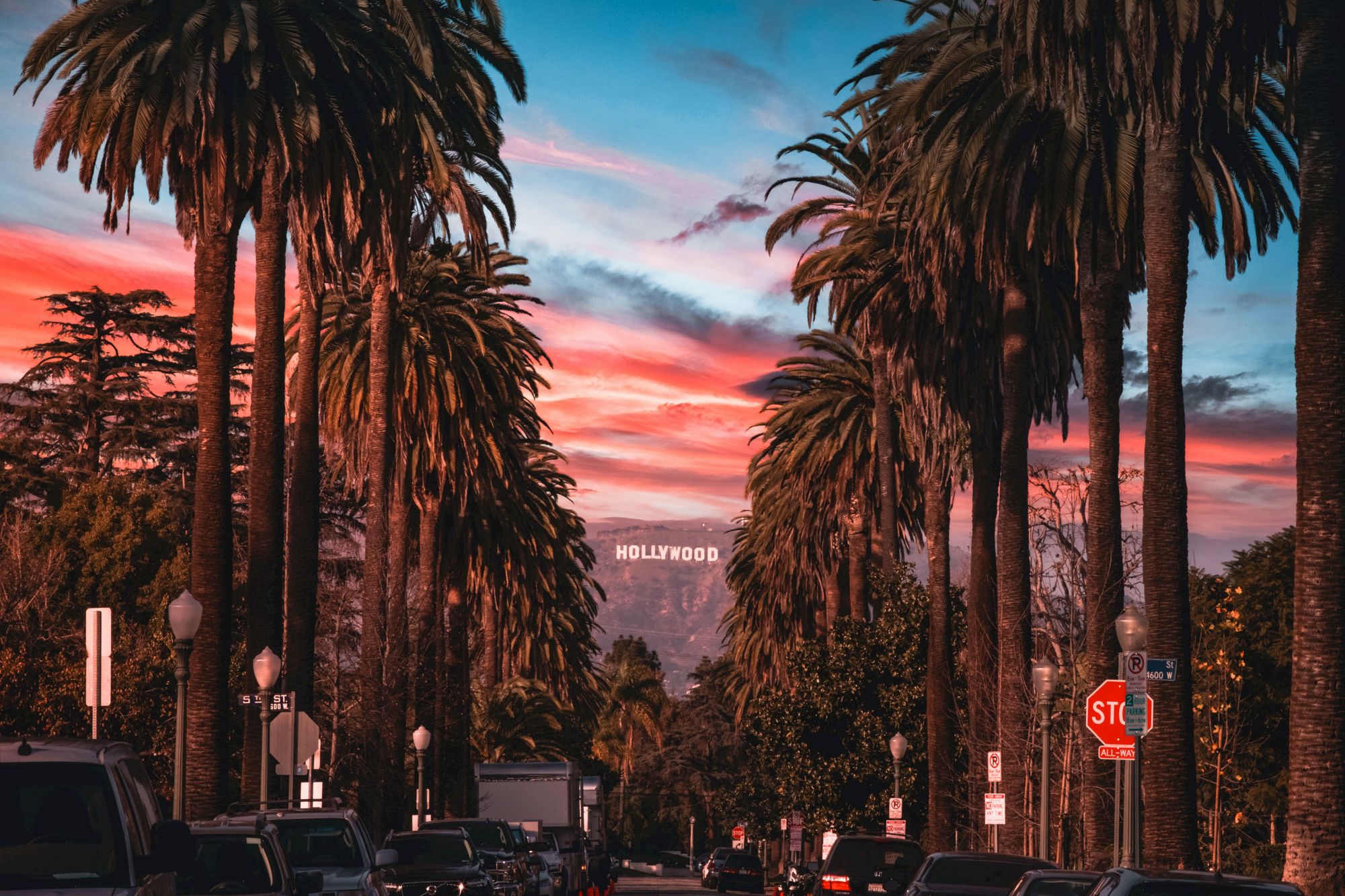 A street lined with tall palm trees at sunset, with the Hollywood sign visible in the background.