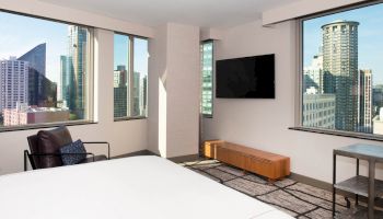 A modern hotel room with large windows showcasing a city view, a wall-mounted TV, a chair, and a wooden table beneath the TV ends the sentence.