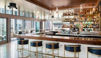 An elegant bar with high stools, a stocked shelf of various bottles, a sleek wooden counter, and large windows opening to a city view.