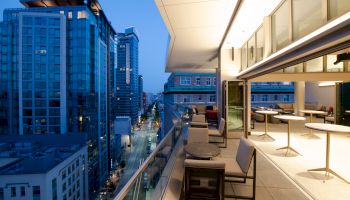 A modern cityscape at dusk with a high-rise apartment balcony, featuring outdoor seating and tables, overlooking the illuminated streets below.