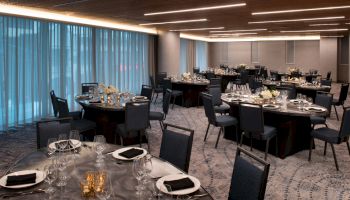 A modern banquet hall is set up with round tables, chairs, silverware, glassware, and floral centerpieces, ready for an event or dinner.