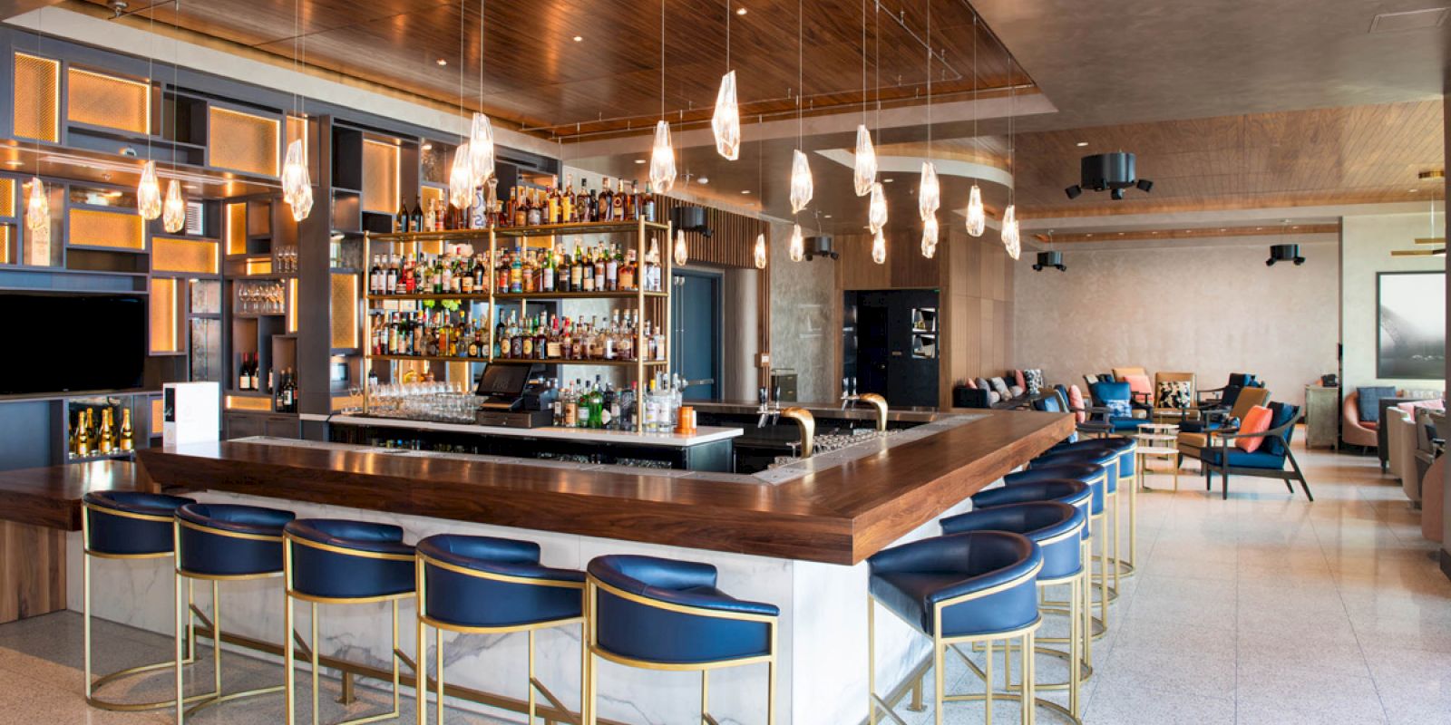 The image depicts a modern bar with a central wooden counter, blue and gold bar stools, a wall of shelves with bottles, and hanging pendant lights.