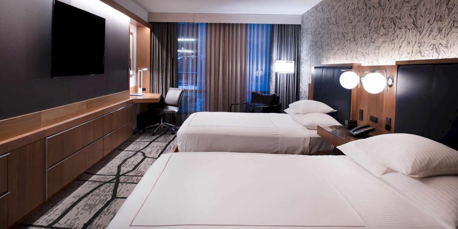 A modern hotel room with two beds, a wall-mounted TV, a desk with a chair, and contemporary decor including a patterned carpet and wallpapered accent wall.