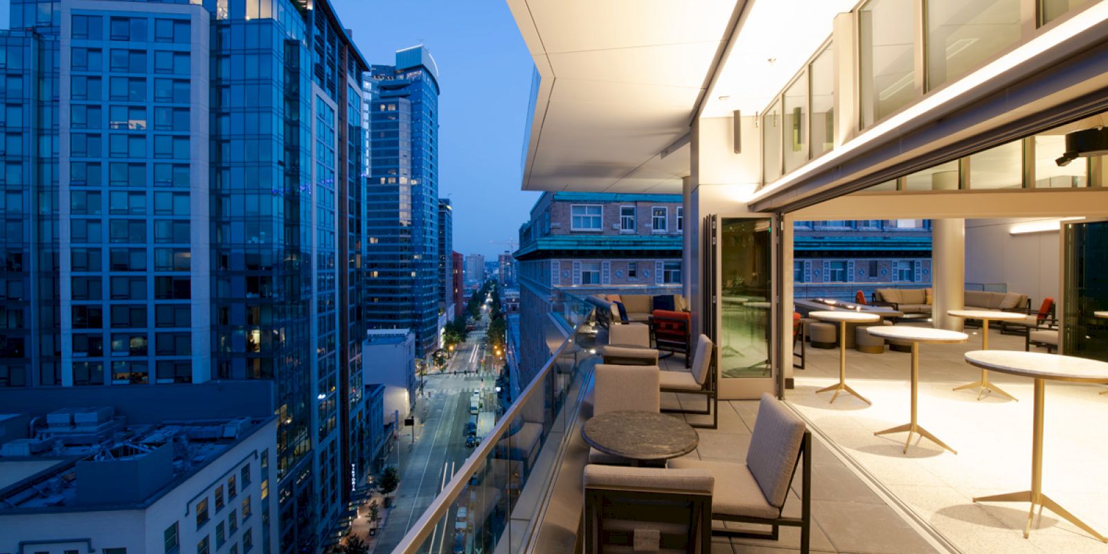 The image shows a modern cityscape at dusk viewed from a high-rise balcony furnished with tables and chairs, overlooking the street below.
