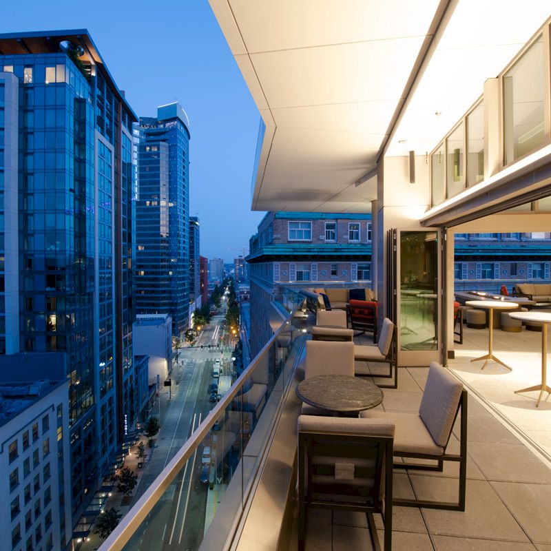 A modern terrace with patio furniture overlooks a city street lined with tall buildings at dusk, providing an elegant urban view.
