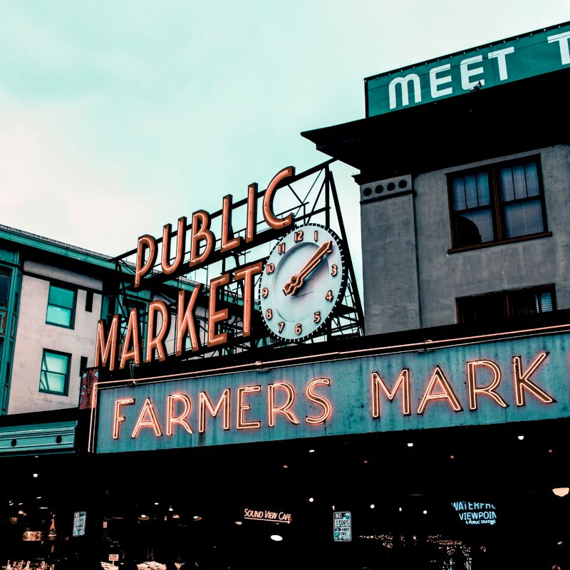 This image shows a "Public Market" sign with a clock above a lit "Farmers Market" sign on a building. The scene appears to be at a market area.