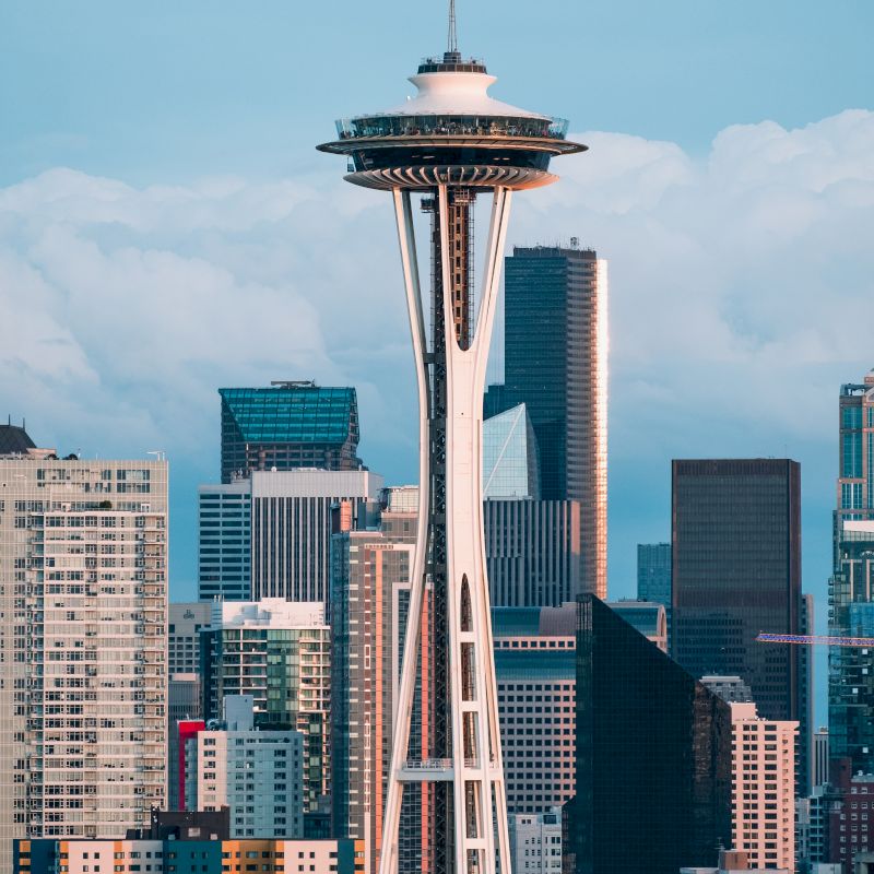 The image shows the Space Needle in Seattle, with the city's skyline in the background, depicting various buildings and the iconic tower against a cloudy sky.