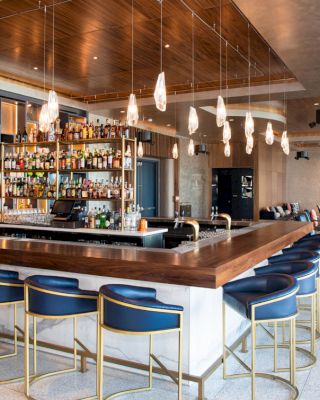 A modern bar with a wooden counter, blue leather bar stools, hanging lights, and a well-stocked bar shelf; seating area in the background.