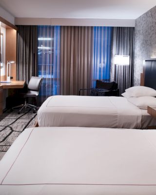 A modern hotel room with two double beds, a wall-mounted TV, a desk, a chair, and a large window with curtains is pictured.
