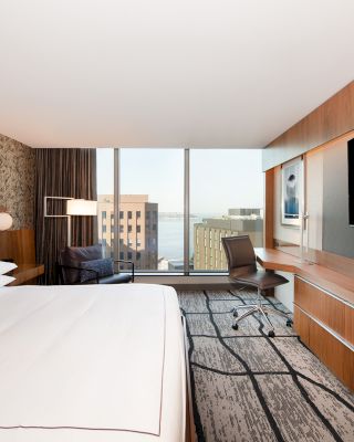 A modern hotel room with a large bed, wall-mounted TV, desk, chair, and window view of buildings and water. The room has contemporary decor.
