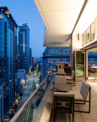 The image shows a modern cityscape at dusk with high-rise buildings and an upscale open-air balcony featuring tables, chairs, and ambient lighting.