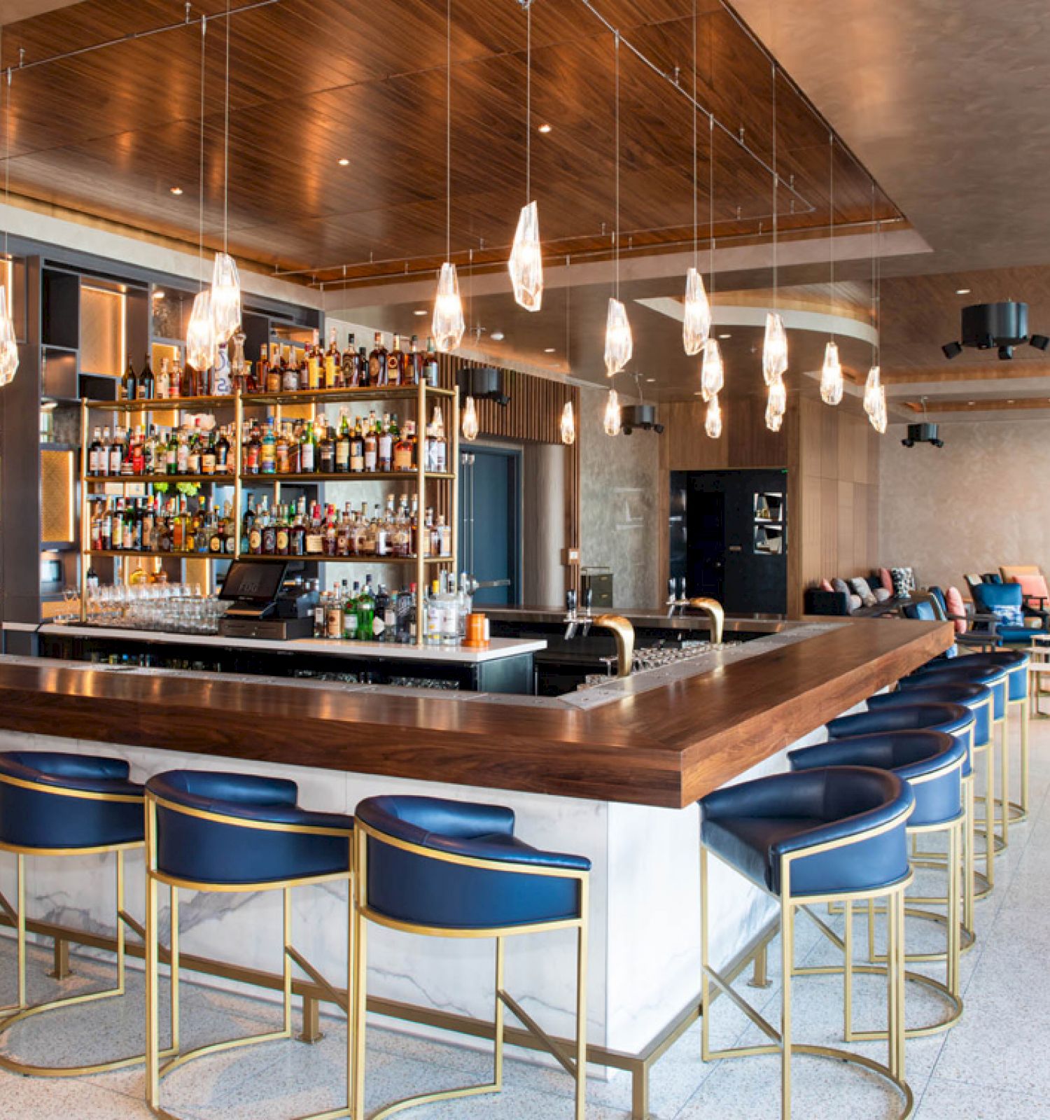 A modern bar with a well-stocked liquor shelf, blue bar stools, pendant lights, and a lounge area in the background ending the sentence.