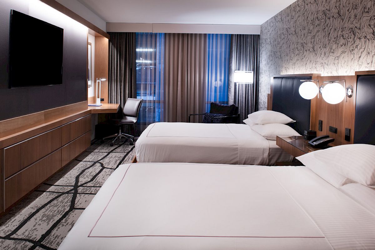 This image shows a modern hotel room with two neatly made beds, a wall-mounted TV, a desk with a chair, and stylish lighting fixtures .