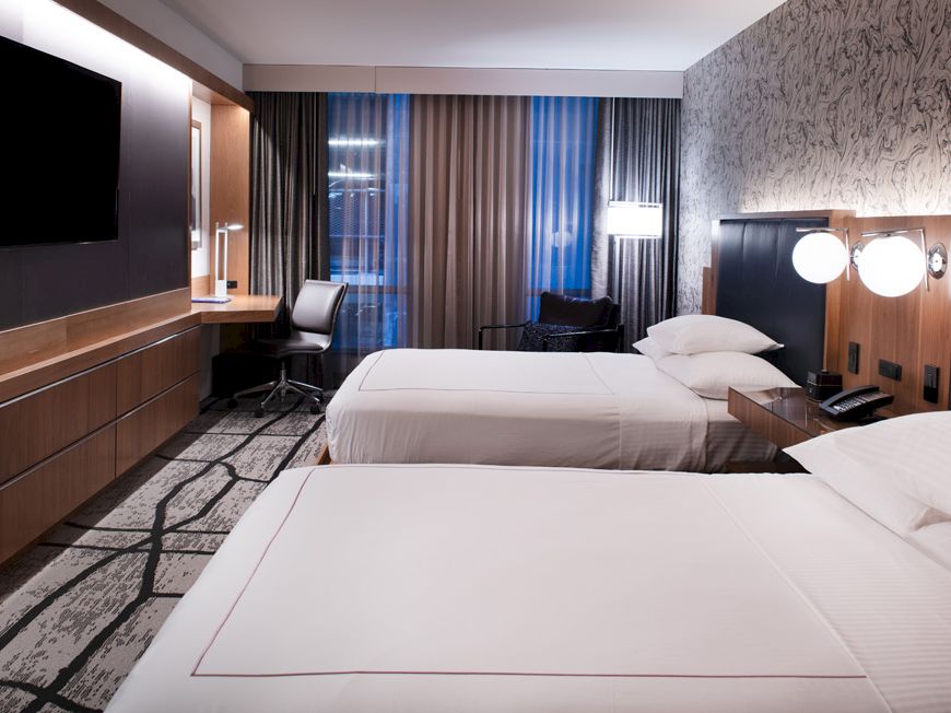 This image shows a modern hotel room with two neatly made beds, a wall-mounted TV, a desk with a chair, and stylish lighting fixtures .