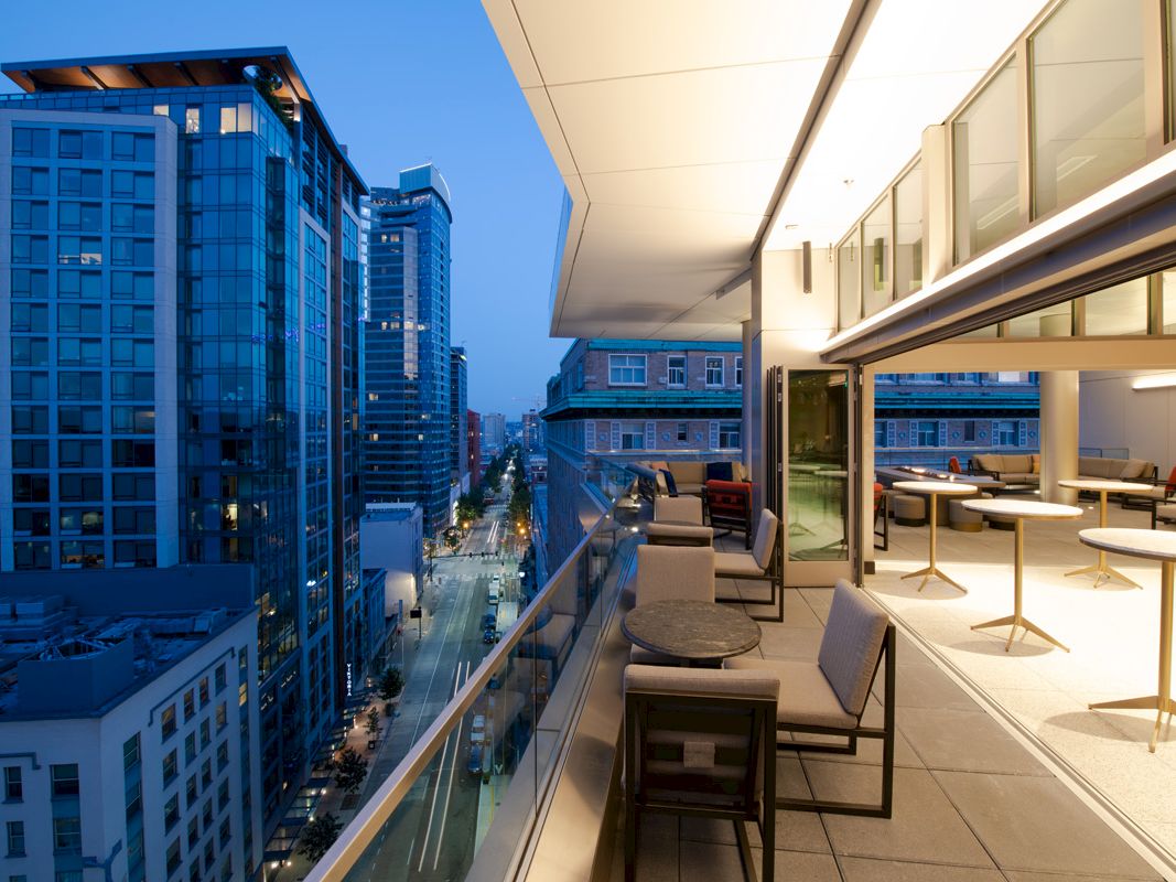 A modern cityscape with high-rise buildings at dusk, featuring a rooftop patio with seating, tables, and a view of the urban street below.
