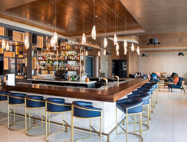 This image shows a modern bar with high chairs, hanging lights, and a well-stocked shelf behind the counter. The setting is elegant and spacious.