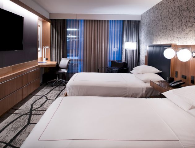 The image shows a modern hotel room with two beds, a wall-mounted TV, a desk, a chair, lamps, and large windows with curtains.