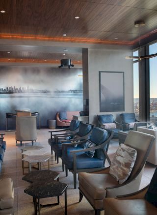 A modern lounge with numerous chairs, tables, and large windows offering a scenic view of a city skyline and waterfront.