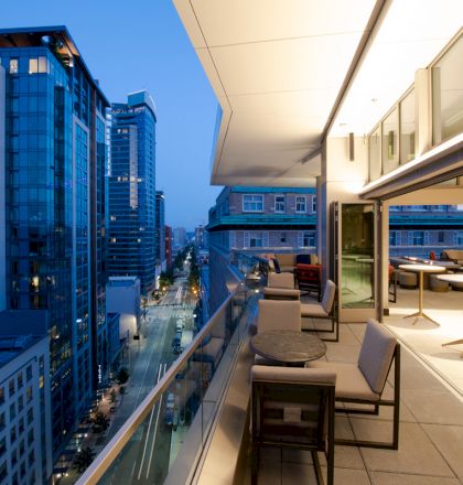 A modern balcony with tables and chairs overlooks a cityscape with tall buildings at dusk, featuring a well-lit, spacious interior area.