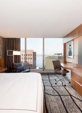 A modern hotel room with a large bed, desk, chair, TV, and window offering a city and water view, featuring contemporary design elements.