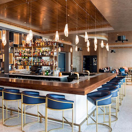 The image shows a modern bar with blue cushioned bar stools, a wooden counter, and a well-stocked shelf of various beverages.