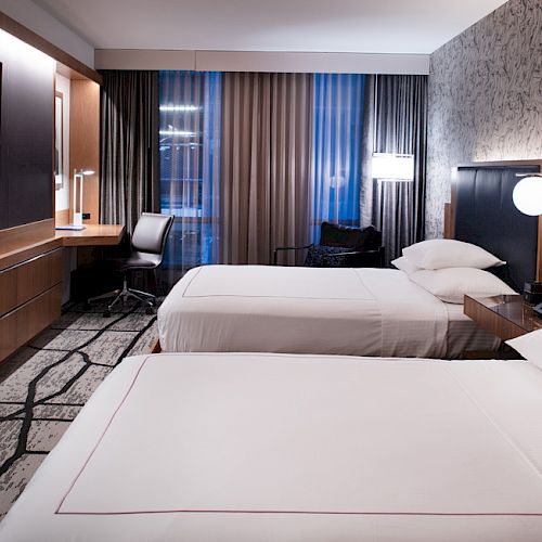 The image depicts a modern hotel room with two neatly made beds, a wall-mounted TV, a desk, and contemporary decor.