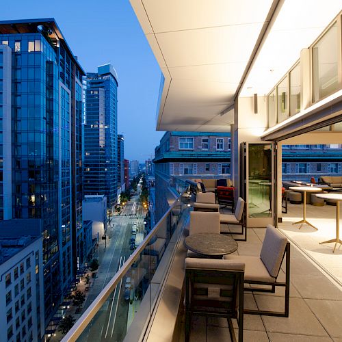 A modern city balcony at twilight, featuring outdoor seating and tables, overlooking a street lined with illuminated high-rise buildings.