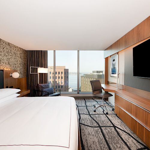 A modern hotel room with a large bed, desk, chair, flat-screen TV, and a window view of city buildings. The decor is contemporary and sleek.