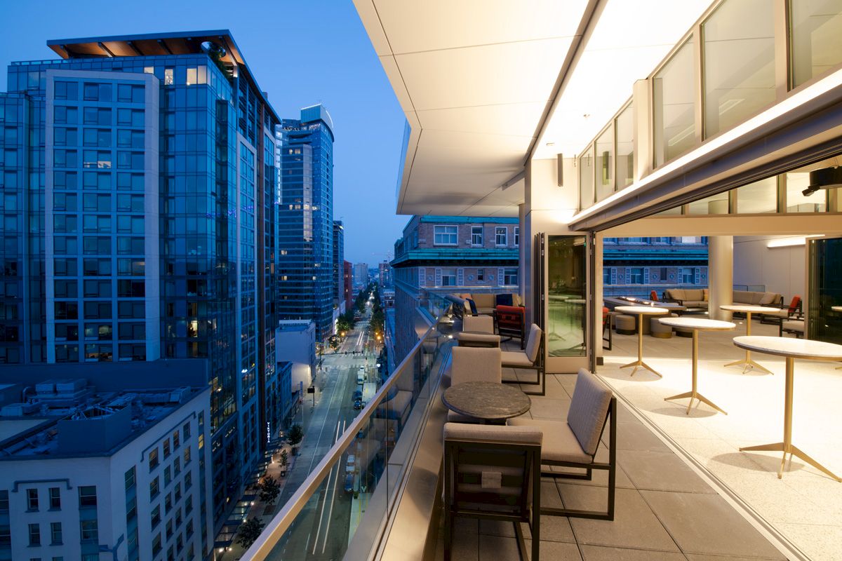 A modern cityscape at dusk with high-rise buildings and a spacious balcony featuring tables, chairs, and an illuminated interior dining area.