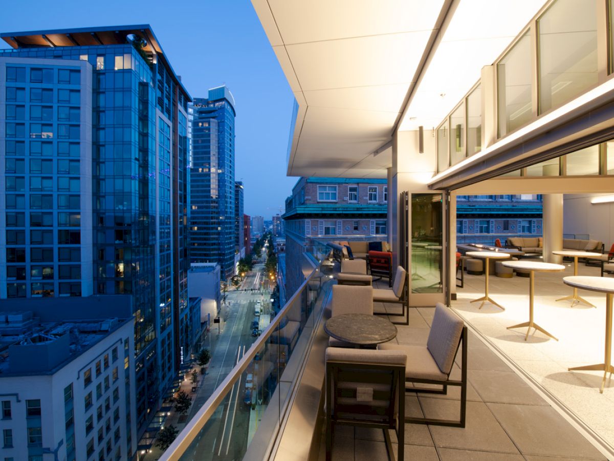 A modern cityscape at dusk with high-rise buildings and a spacious balcony featuring tables, chairs, and an illuminated interior dining area.