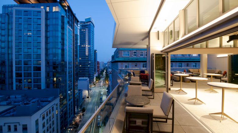 A modern cityscape at dusk with high-rise buildings and a spacious balcony featuring tables, chairs, and an illuminated interior dining area.