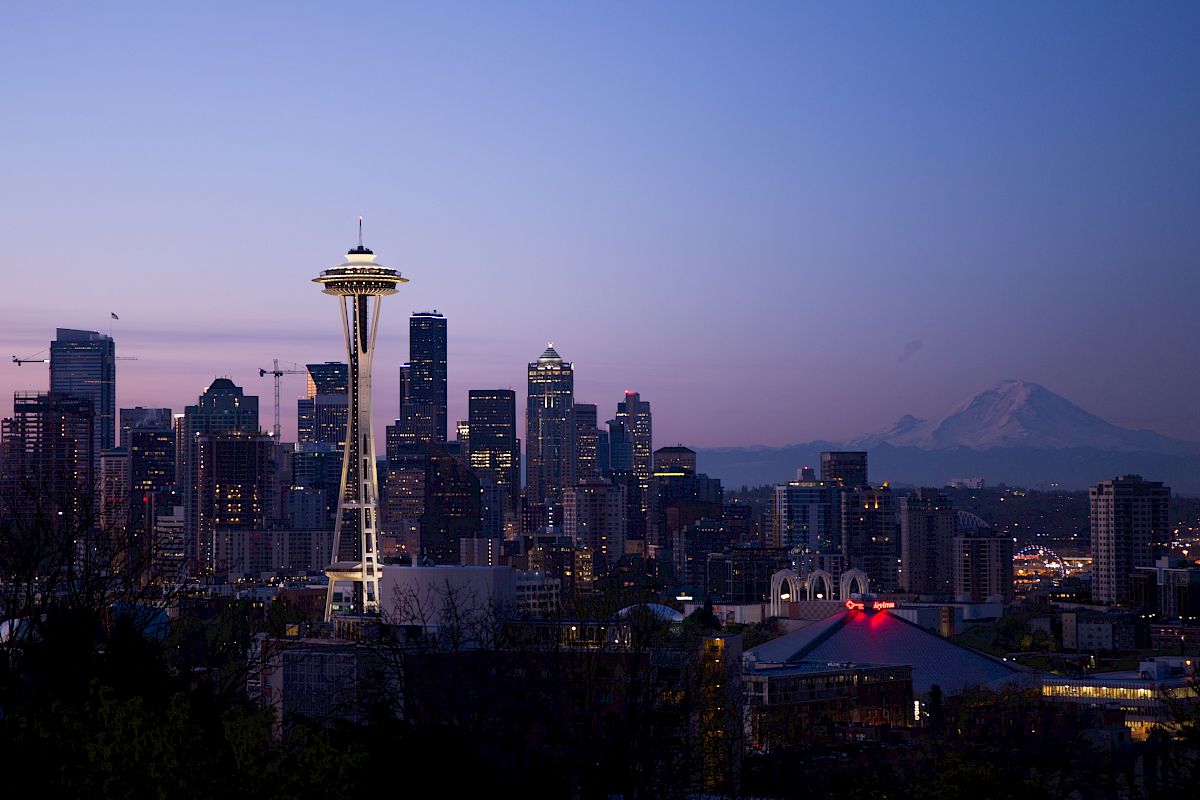 This image shows the Seattle skyline at dusk, featuring the Space Needle and surrounding high-rise buildings, with Mount Rainier visible in the background.