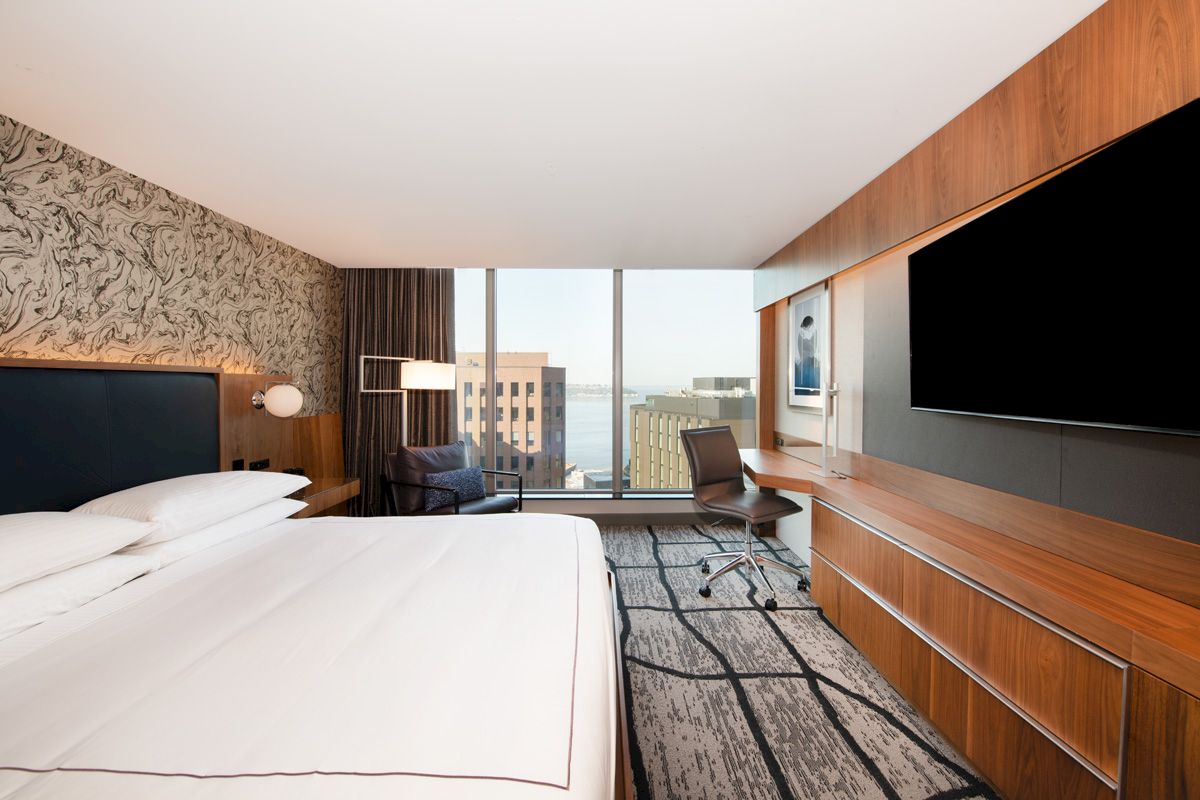 A modern hotel room with a large bed, a wall-mounted TV, a desk with a chair, and a window offering a view of city buildings and water.