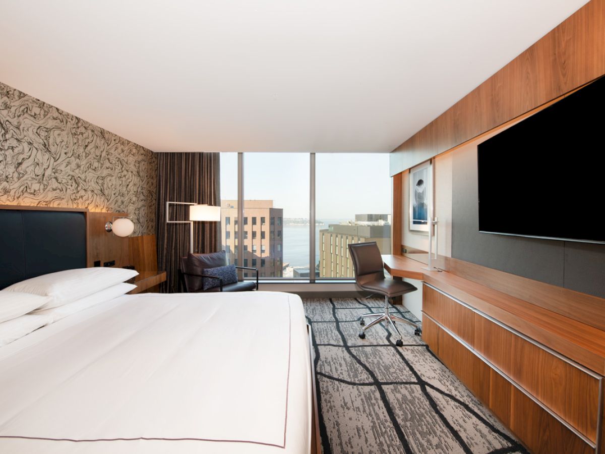 A modern hotel room with a large bed, a wall-mounted TV, a desk with a chair, and a window offering a view of city buildings and water.